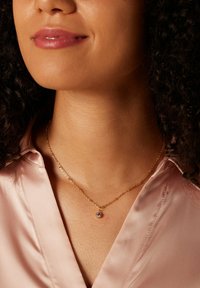 Gouden ketting met een cirkelvormige hanger met een paarse edelsteen, gedragen over een lichtroze satijnen blouse.