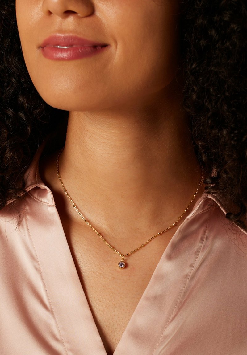 Gouden ketting met een cirkelvormige hanger met een paarse edelsteen, gedragen over een lichtroze satijnen blouse.