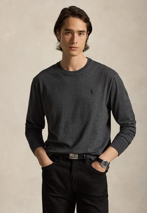 Jeune homme aux cheveux foncés porte une chemise gris foncé à manches longues et un pantalon noir, debout, les mains dans les poches, devant un fond neutre.