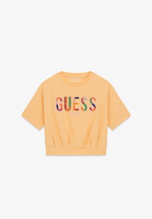 T-shirt court de couleur pêche avec le texte multicolore "GUESS" et "EST. 1981" imprimés sur le devant.