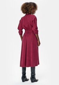 Kobieta o kręconych włosach ubrana w bordową sukienkę z długim rękawem, sięgającą kolan, z marszczonym pasem i czarne kozaki za kolano, zwrócona tyłem.