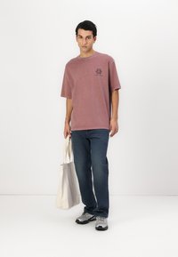 Bawełniany t-shirt w zgaszonym mauve z okrągłym dekoltem i krótkimi rękawami, z ciemną grafiką po lewej stronie klatki piersiowej; w połączeniu z niebieskimi dżinsami i beżową torbą.