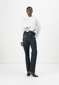 White hoodie with "Karl" printed in black, paired with dark blue jeans featuring front pockets and gold button accents. Black heeled shoes.