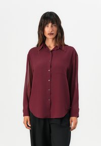 Camicia bordeaux con bottone, maniche lunghe trasparenti, tasca sul petto, orlo arrotondato e finiture in argento discreto. Abbinata a pantaloni neri.