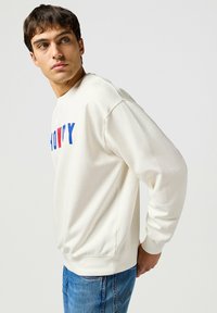 Sudadera de color crema con el texto "HOWDY" en rojo y azul, de mangas largas y puños acanalados. Combinada con pantalones vaqueros de mezclilla azul.