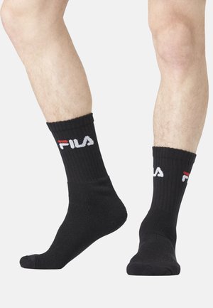 Jambes portant des chaussettes noires côtelées avec le logo FILA blanc et rouge, un pied pointé, l'autre à plat sur un fond clair.