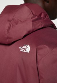 Karmelowa kurtka z kapturem wykonana z teksturowanej tkaniny, z białym logo na górnej lewej stronie. Zamek błyskawiczny oraz widoczne szwy boczne.