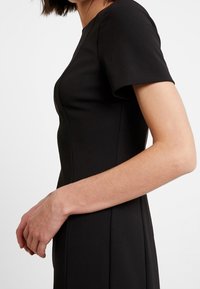 Schwarzes, tailliertes Kleid mit kurzen Ärmeln, das eine glatte Textur und Nahtdetails aufweist. Die Silhouette ist so zugeschnitten, dass sie eine geschmeidige Erscheinung hat.