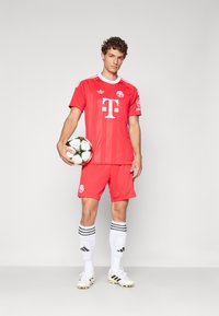 Rode voetbalshirt met witte accenten, voorzien van een logo. De speler houdt een zwart-wit voetbal vast en draagt bijpassende rode shorts en witte sokken.