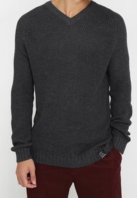 Mann in einem dunkelgrauen, gestrickten V-Ausschnitt-Pullover und dunkelroten Hosen, mit einer Hand in der Tasche vor einem schlichten Hintergrund.