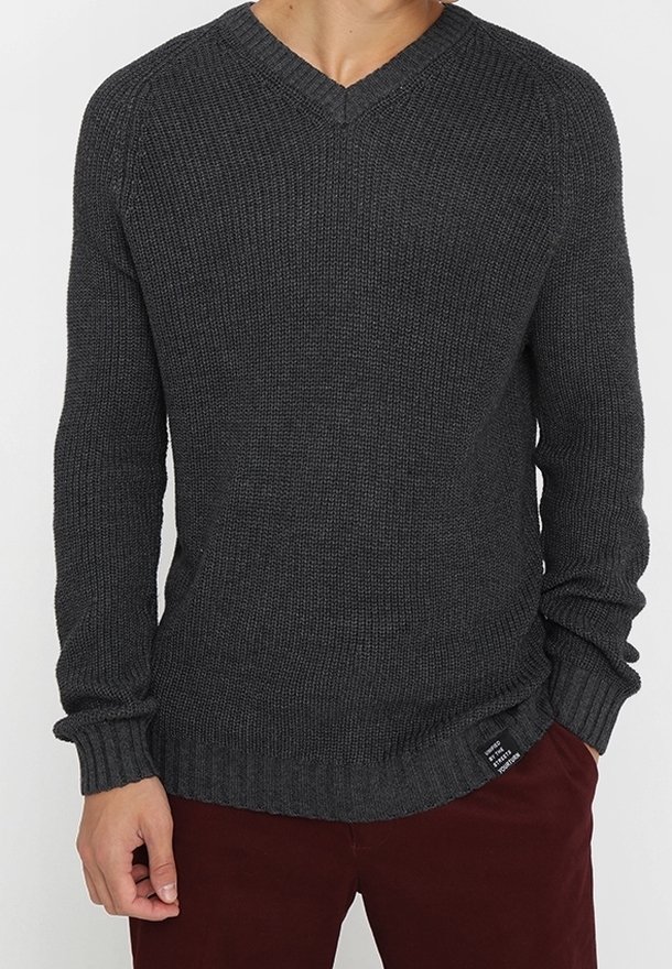 Mann in einem dunkelgrauen, gestrickten V-Ausschnitt-Pullover und dunkelroten Hosen, mit einer Hand in der Tasche vor einem schlichten Hintergrund.