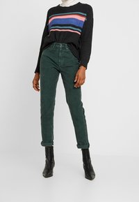 Person som bär en svart tröja med färgglada ränder, gröna uppvikta kordbyxor och svarta ankelstövlar som står mot en enfärgad bakgrund.