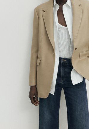 Personne portant un blazer beige sur une chemise blanche à boutons et un haut en maille blanc, associé à un jean bleu foncé, sur un fond uni.