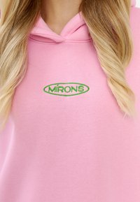 Felpa rosa realizzata in tessuto morbido con tasca frontale. Presenta un logo ovale verde con la scritta "MÍRONS" sul petto e un cappuccio con stringa.