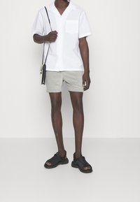 Camisa blanca de manga corta con sutiles rayas, pantalones cortos grises y sandalias negras con correas ajustables. Incluye un pequeño bolsillo frontal.