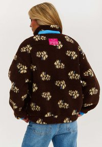 Hnědá fleecová bunda s modrou podšívkou, zdobená květinovým vzorem v krémové a modré barvě. Na zadním límci je růžová etiketa.