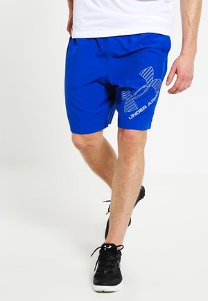 Mann, der leuchtend blaue Under Armour Shorts und schwarze Sportschuhe trägt und vor einem weißen Hintergrund geht.