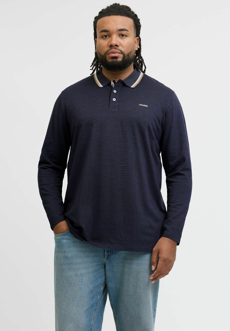Mann mit Bart und Dreadlocks, der ein marineblaues Langarm-Poloshirt und helle Jeans trägt, steht mit einer Hand in der Tasche vor einem weißen Hintergrund.