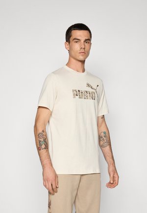 Mand iført en beige Puma T-shirt og matchende bukser, som viser tatoveringer på begge underarme, står foran en ensfarvet lys baggrund.