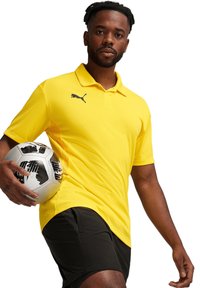 Geel poloshirt met een gestructureerd patroon, zwart logo op de borst, gecombineerd met zwarte shorts, terwijl hij een patroonvoetbal vasthoudt.