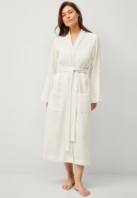 Peignoir de bain blanc en tissu à motif gaufré, avec des manches longues, un design enveloppant, une ceinture à nouer et deux poches avant, s'étendant jusqu'à mi-mollet.