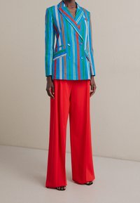 Gestreepte blauwe en groene blazer met rode accenten, gecombineerd met rode, wijde broekspijpen. De blazer heeft twee zakken en een dubbelgeknoopt ontwerp.