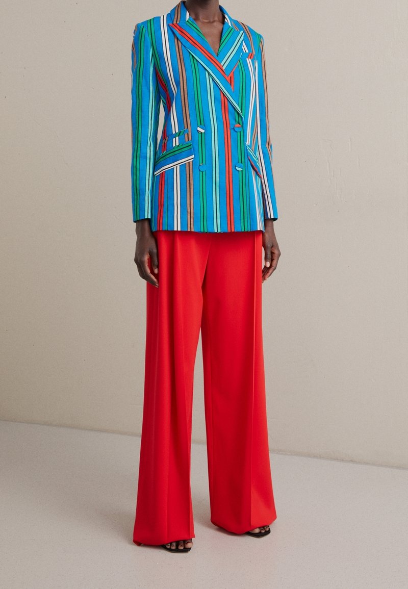 Gestreifter blau-grüner Blazer mit roten Akzenten, kombiniert mit roten, weit geschnittenen Hosen. Der Blazer verfügt über zwei Taschen und ein doppeltreihiges Design.