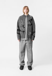 Cardigan de punto gris con patrón abstracto, combinado con una camiseta gris clara y pantalones anchos grises, con un detalle en cadena. Calzado negro.