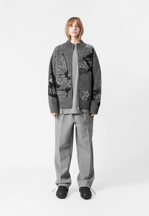 Cardigan de punto gris con patrón abstracto, combinado con una camiseta gris clara y pantalones anchos grises, con un detalle en cadena. Calzado negro.