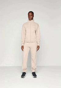 Beżowa kurtka zapinana na zamek z wysokim kołnierzem i pasującymi dresami, wykonana z miękkiego materiału. Posiada ściągacze na rękawach i czarne buty sportowe.