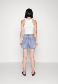 Top sin mangas blanco combinado con pantalones cortos de mezclilla azul claro. Los pantalones cortos tienen dos bolsillos traseros y un dobladillo deshilachado. La modelo se encuentra sobre un fondo liso.