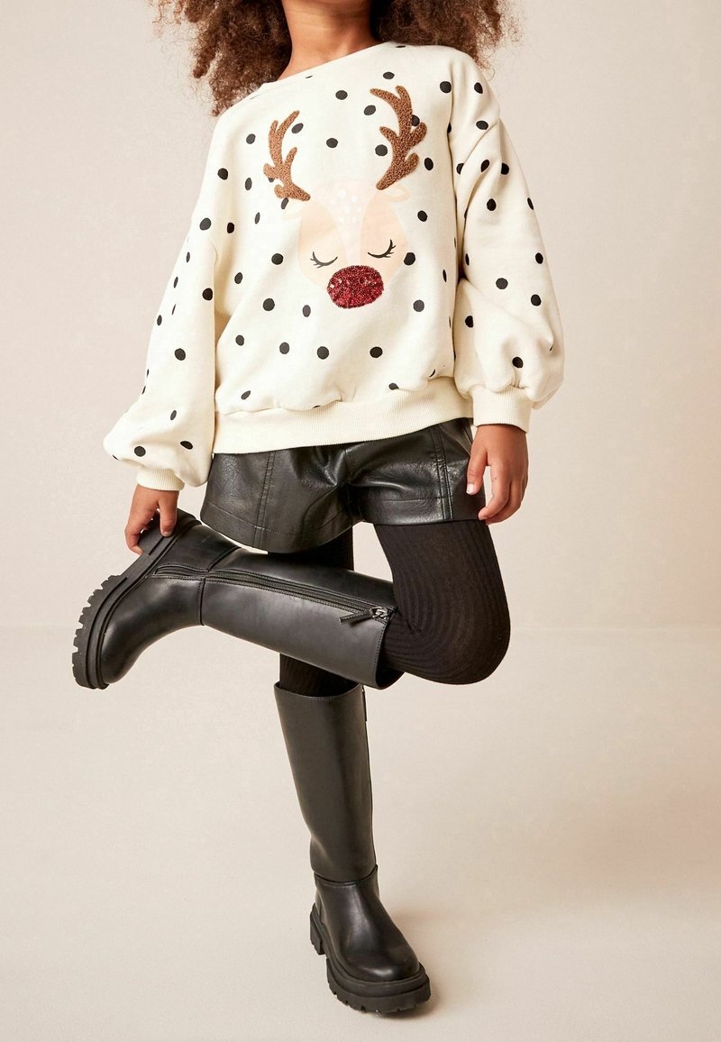 Enfant portant un pull blanc à pois avec un motif de renne, un short en cuir noir, des collants noirs et des bottes noires montantes, levant une jambe.