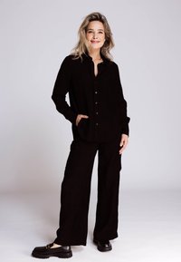 Schwarzes Cordhemd mit Knopfleiste und Kragen, kombiniert mit passenden locker sitzenden Hosen. Schwarze, klobige Loafers vervollständigen das Outfit.