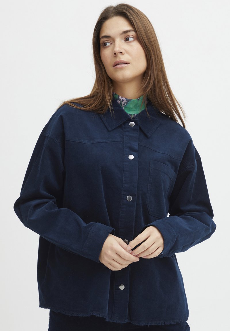 Giacca camicia in corduroy blu navy con chiusura a bottoni, colletto a punta e taschino sul petto. Presenta un orlo sfilacciato e maniche lunghe.