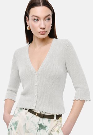 Donna che indossa un cardigan a coste grigio chiaro con bordi smerlati e bottoni bianchi, abbinato a pantaloni a fantasia floreale e orecchini dorati.