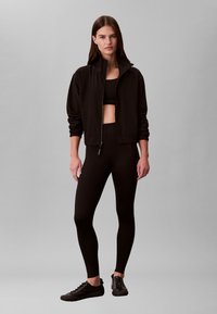 Sieviete stāv, tērpusies melnā rāvējslēdzēja džemperī, melnā apgrieztā topiņā, melnās legingās un melnās sporta apavos pret pelēku fonu.