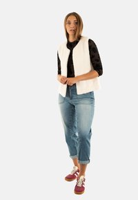 Gilet bianco a maniche corte con texture sopra una camicia nera a maniche lunghe con fantasia, abbinato a jeans in denim azzurro chiaro e sneakers bordeaux.