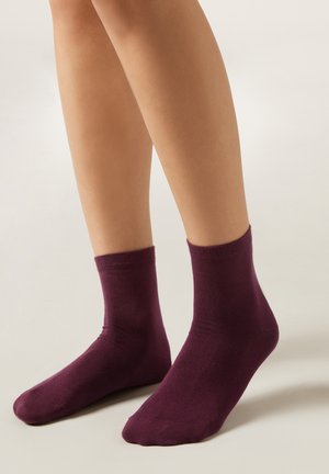 Gambe con calzini viola scuro in cotone alla caviglia, in piedi su una superficie chiara con uno sfondo neutro.