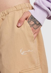 Beige stoffshorts med elastisk midje og sidelomme, med en hvit brodert logo-detall. Teksturen ser jevn og lett ut.