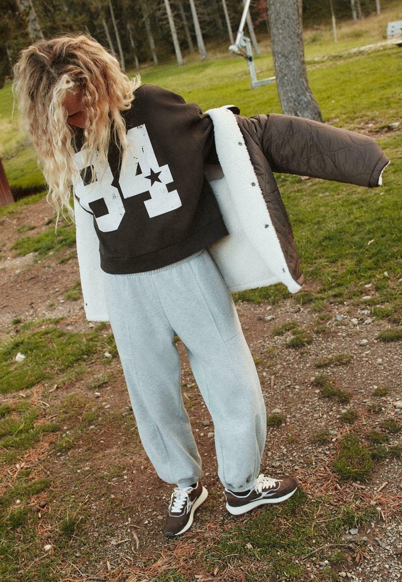 Pantaloni grigi in felpa abbinati a una felpa corta nera con una grafica bianca "84" e una giacca imbottita marrone con un interno bianco.