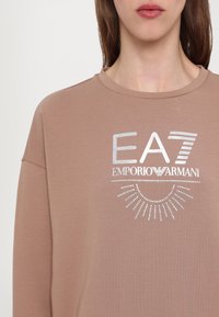Barna kapucnis pulóver, amelyen fényvisszaverő EA7 Emporio Armani logó található napfény-szerű mintával. Az anyag lágyan puha; bordás kerek nyakkivágás.