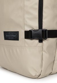 Beige Eastpak-ryggsäck med en texturerad finish, utrustad med en svart dragkedja, svarta remspännen och en svart logotyplapp.
