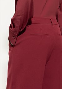 Burgunderfarbene Bluse mit glatter Textur und umgeschlagenen Ärmeln, kombiniert mit passenden, taillierten Hosen mit einem eleganten Design und dezenten Gesäßtaschen.