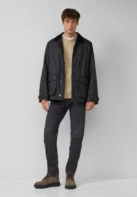 Zwarte, waterdichte jas met een kraag van schapenvacht, knoopsluiting en twee voorzakken, gedragen over een beige sweater en donkere jeans.