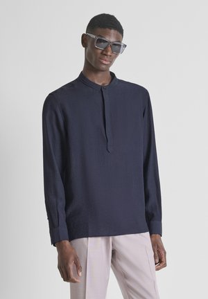 Uomo che indossa una camicia blu scuro a maniche lunghe con colletto alla coreana e pantaloni beige chiaro, posando davanti a uno sfondo chiaro e semplice.