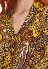 Tessuto con motivo paisley giallo e marrone, caratterizzato da intricate spirali e disegni floreali, con scollatura a V e texture morbida.