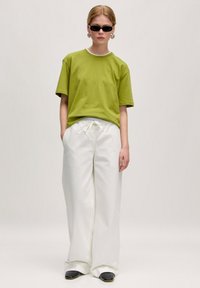 Grünes Rundhals-T-Shirt mit kurzen Ärmeln, weiße weite Hose mit Schnürung und schwarze Sonnenbrille. Glatter Stoff und lockere Passform.