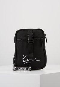 Borsa a tracolla nera in tessuto con chiusura a zip, dotata di dettagli bianchi con firma e di una tracolla regolabile a fantasia. Design rettangolare compatto.