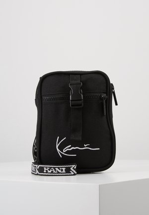 Schwarze Stoff-Crossbody-Tasche mit Reißverschluss, ausgestattet mit weißen Signaturdetails und einem gemusterten verstellbaren Gurt. Kompakte rechteckige Form.