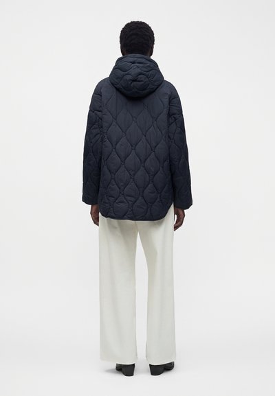 Personne debout, de dos, portant une veste matelassée sombre à capuche et un pantalon blanc à jambes larges, sur un fond clair uni.